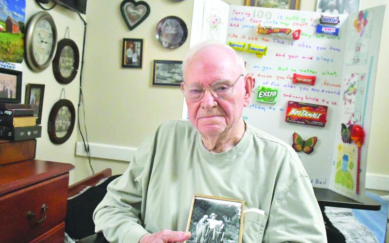 Megan Broome/The Clayton Tribune. Ervin Chastain enjoys smiling, laughing and telling stories of his adventures in life. He was born on June 10, 1923, and recently celebrated the milestone with family, friends and former coworkers at Battle Branch Baptist Church. Pictured here, he holds a photo of himself at age 20 or 21 with parents Mack and Rosie Chastain. Behind him is a poster made by family members to honor his 100 years. 