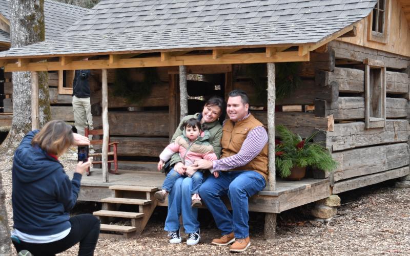 Enoch Autry/The Clayton Tribune. With Foxfire volunteer Laura West taking a family photo, Natalie Strasser and John Strasser pose with their 2-year-old daughter Savannah as they sit on the porch of the Foxfire miniature house, ironically, named the Savannah Cabin in the Children’s Village during the Dec. 16 “A Foxfire Christmas” event. Left: Kim Cannon from the Rabun County Public Library read a variety of books that depicted pioneer life in the Appalachian Mountains on the Shooting Creek Cabin porch.