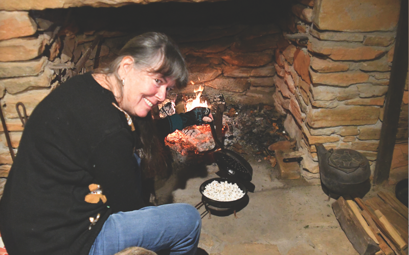 Enoch Autry/The Clayton Tribune. Foxfire volunteer Cathy Stiles heats up corn kernels over hot embers to make popcorn at the Dec. 16 “A Foxfire Christmas” in Mountain City. The popcorn was strung on string around the Christmas tree and, yes, some of the popcorn also was eaten. Left: Kim Cannon from the Rabun County Public Library read a variety of books that depicted pioneer life in the Appalachian Mountains on the Shooting Creek Cabin porch.