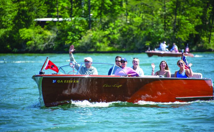Submitted photo. A boat owner waves to the crowd during a previous LBCA Wooden Boat Parade show on Lake Burton. Left: Photo courtesy Conner Horn.  Rabun County is the Wooden Boat Capital of Georgia and Lake Burton Wooden Boat owners hold up the sign at the May 12 Captain’s reception. 