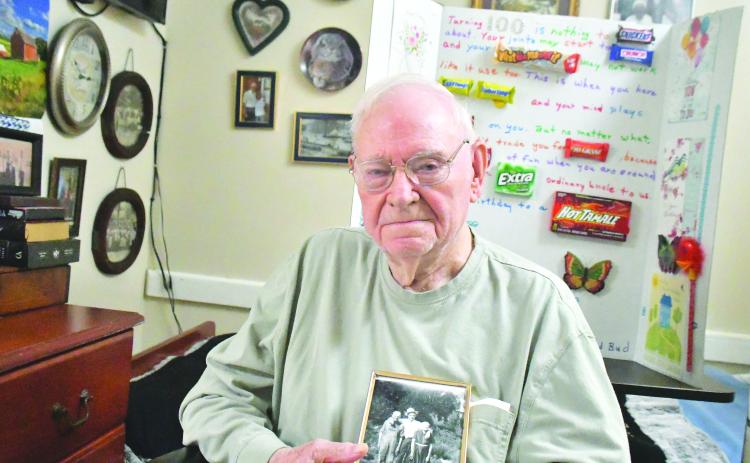 Megan Broome/The Clayton Tribune. Ervin Chastain enjoys smiling, laughing and telling stories of his adventures in life. He was born on June 10, 1923, and recently celebrated the milestone with family, friends and former coworkers at Battle Branch Baptist Church. Pictured here, he holds a photo of himself at age 20 or 21 with parents Mack and Rosie Chastain. Behind him is a poster made by family members to honor his 100 years. 