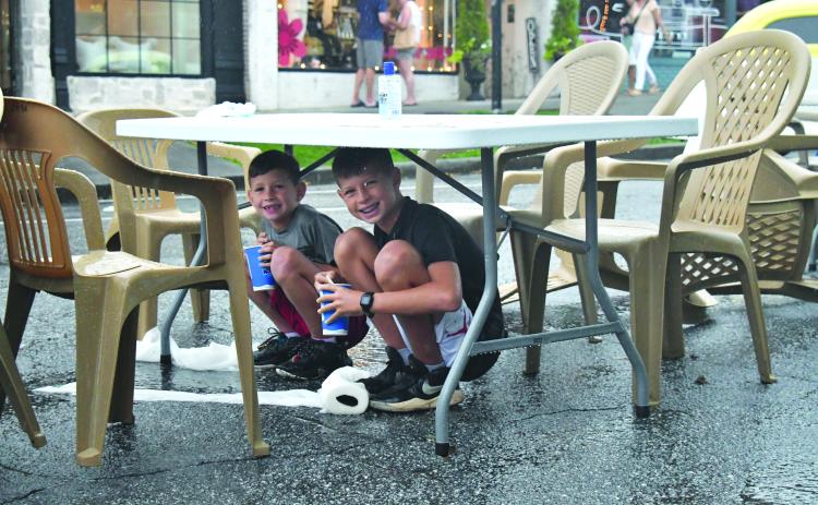 Enoch Autry/The Clayton Tribune. With their cups they got from a food truck, 11-year-old Grayson Miller and his brother Asher, 8, have a reason to smile as they stay dry under a table at the Downtown Clayton Block Party as a storm blew through Main Street on Aug. 12. Water droplets can be seen coming off the table as the paper towels next to their feet on the street are saturated. 