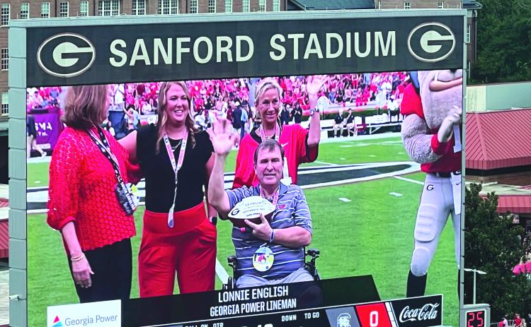 Submitted. Georgia Power lineman and Clayton resident Lonnie English, waves to over 96,000 fans while being honored at the Sept. 16 Georgia vs South Carolina football game after overcoming a traumatic leg injury. Joining English on the 50-yard line of Dooley Field at Sanford Stadium. Pictured from left to right is GA Power Chairman, President and CEO, Kim Greene, English’s daughter Logan, English’s wife Caroline and Georgia Mascot Hairy Dawg. 