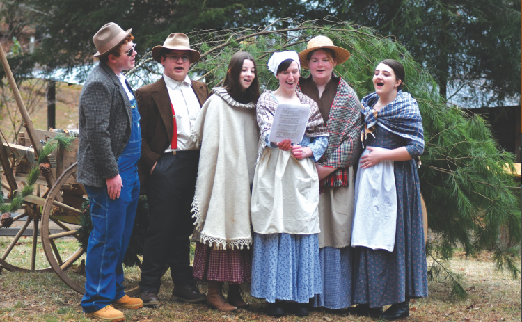 Enoch Autry/The Clayton Tribune. Rabun County High School students Isaac Craig, Andy Leff, Isabel Craine, Leizle Mason, Sarabeth Speed and Marlena Turpin sing holiday songs near the Warwoman Cabin and Phillips Cabin at the Dec. 16 annual “A Foxfire Christmas” in Mountain City. Left: Kim Cannon from the Rabun County Public Library read a variety of books that depicted pioneer life in the Appalachian Mountains on the Shooting Creek Cabin porch.